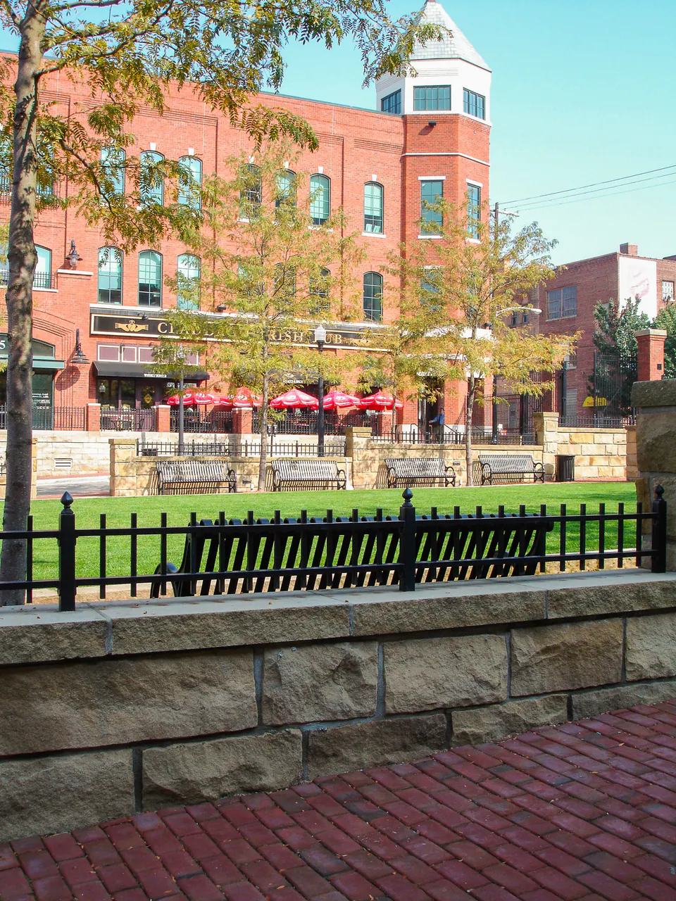 View through black railing toward courtyard lawn, brick buildings, seating umbrellas, and trees.